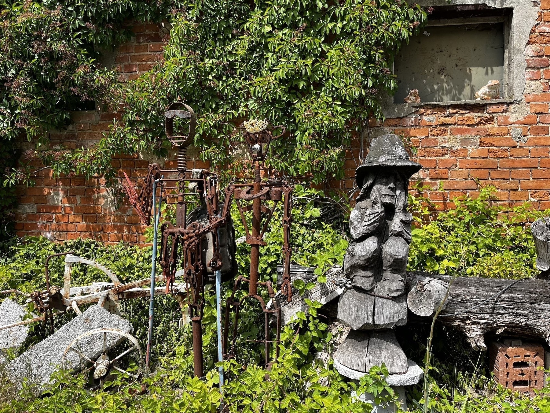 Eine verfallene, überwucherte Mauer umrahmt skurrile Metallskulpturen und eine Holzfigur im Freien.
