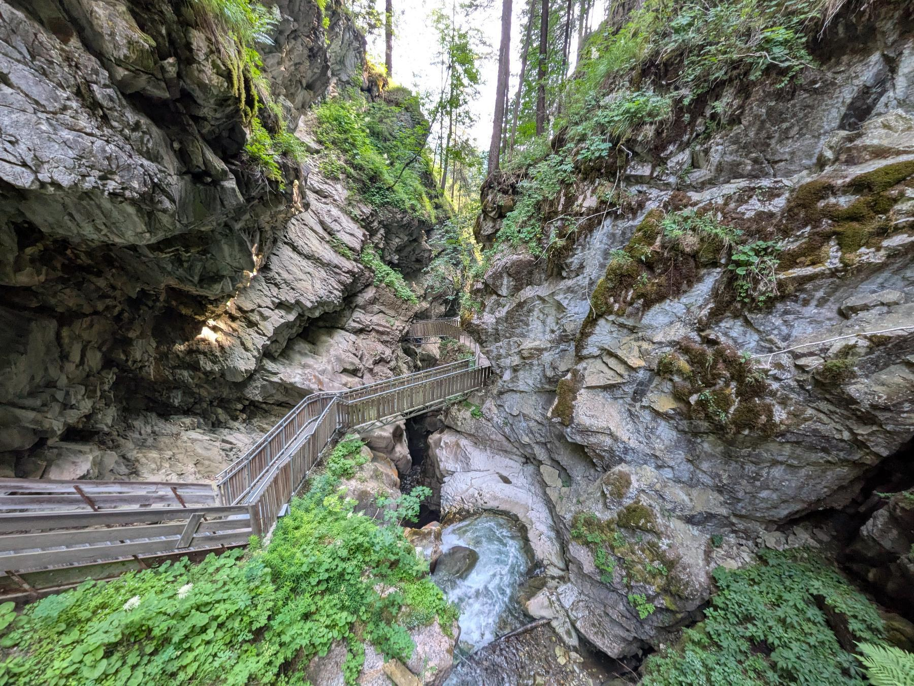 Ein hölzerner Steg führt durch eine enge, felsige Schlucht (Gilfenklamm) mit fließendem Wasser und umgeben von dichter Vegetation.