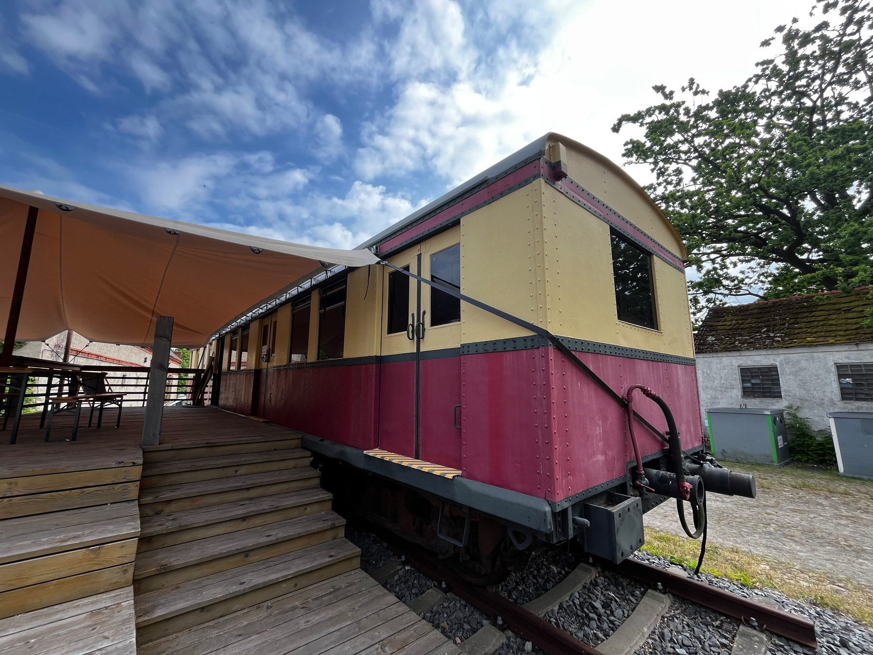 Ein alter, rot-beiger Eisenbahnwaggon steht auf Schienen neben einer überdachten Terrasse unter blauem Himmel.