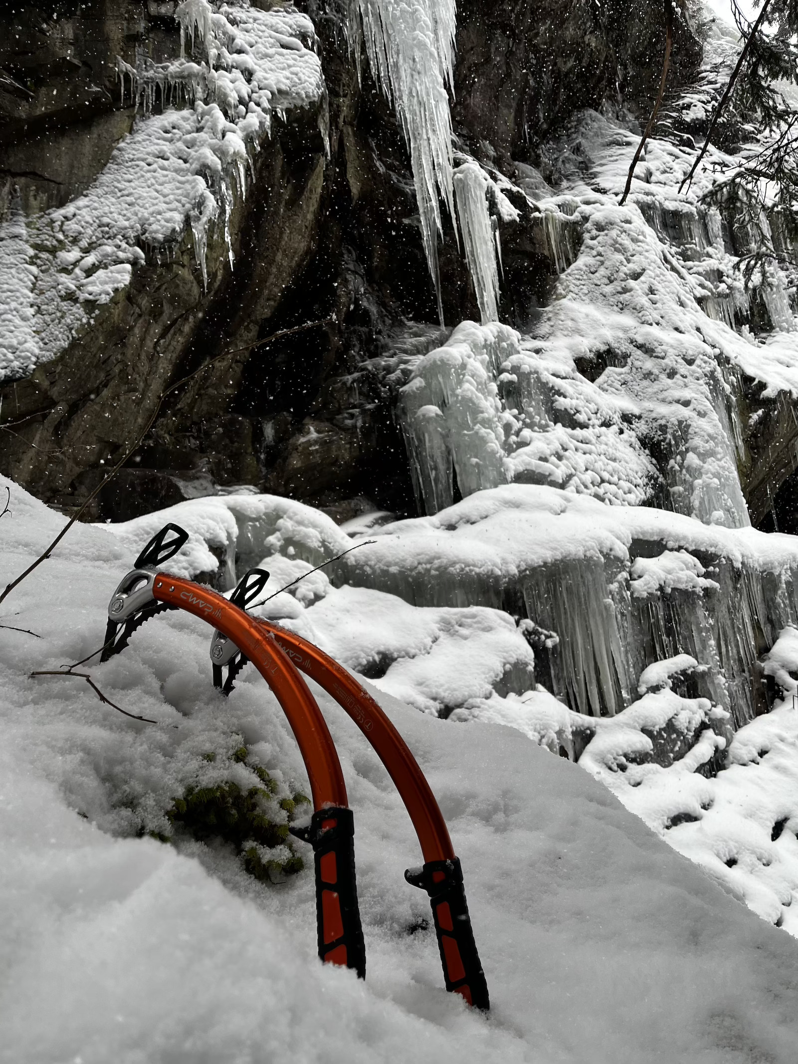 Zwei orangefarbene Eispickel stecken im Schnee vor einer eisbedeckten Felswand mit großen Eiszapfen.