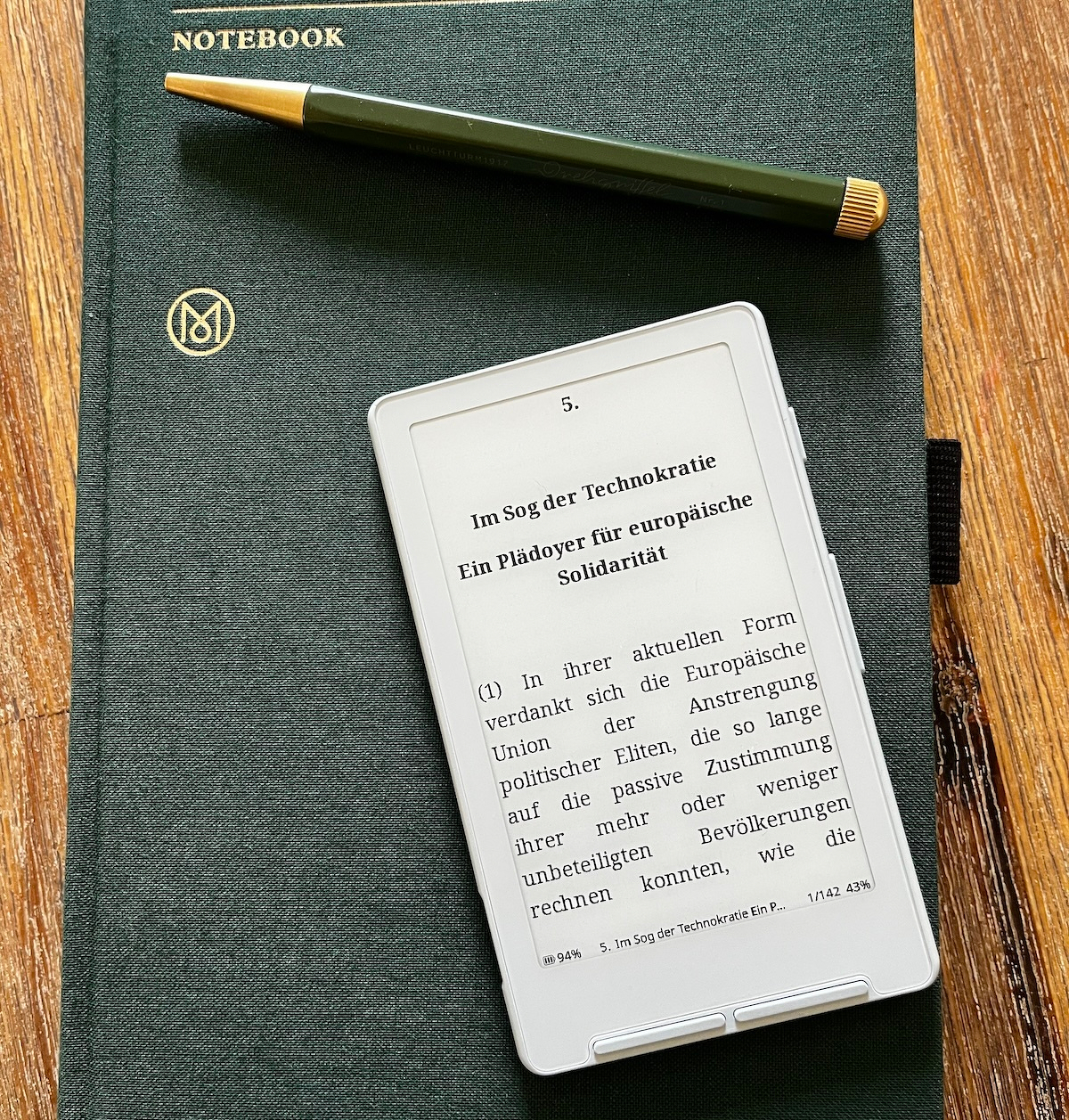 Ein grünes Notizbuch, ein Stift und ein E-Reader liegen auf einem Tisch.