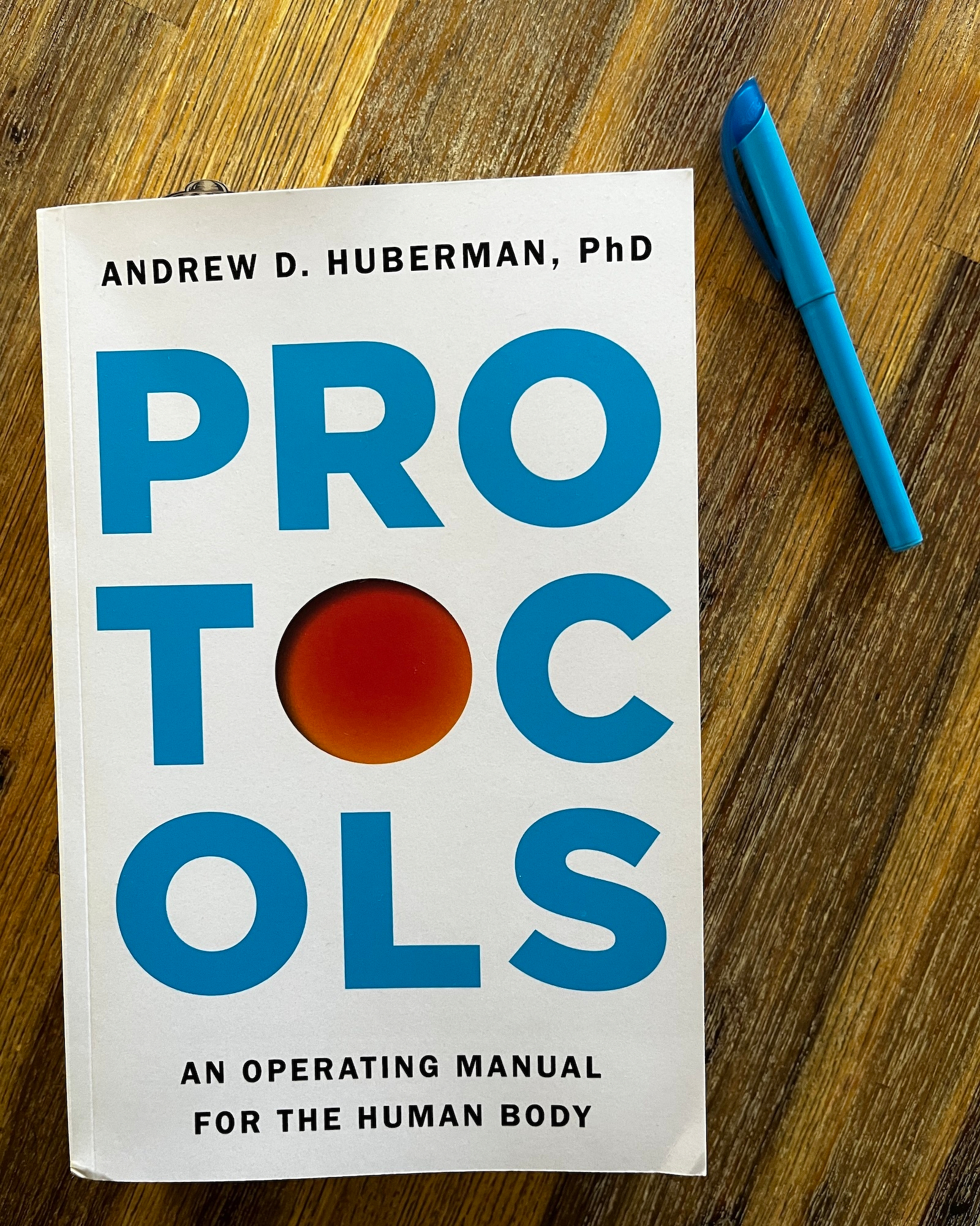 Ein Buch mit dem Titel „Protocols“ von Andrew D. Huberman, PhD, liegt auf einer Holzoberfläche neben einem blauen Stift.